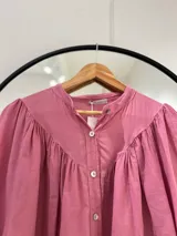 Camisa rosa de manga larga con puños, cuello mao y cierre frontal con botones. Tiene corte y frunce en la parte delantera.