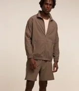 Campera deportiva color marrón, con cuello alto, cierre frontal con cremallera, manga larga, bolsillos laterales y detalles de etiqueta aplicada y rayas.
