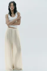 Pantalón palazzo color crudo, de tiro alto, confeccionado en lyocell. Presenta bolsillos laterales ocultos, detalle de pliegues en el delantero y cierre frontal con cremallera y botones.