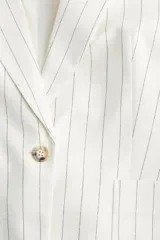 Blazer entallado color blanco con finas rayas verticales negras, solapas de pico, cierre de un botón, bolsillos delanteros de parche y puños con abertura y botón. Forrado.