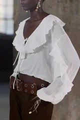 Blusa color marrón con escote en pico, manga larga abullonada con elástico en los puños y volantes en el escote y mangas.