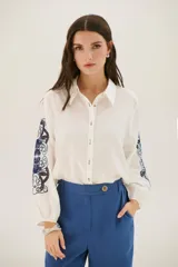 Camisa blanca de mangas largas con puños elásticos, cuello clásico y cierre frontal con botones. Presenta bordados florales en tonos azules y marrones en las mangas.