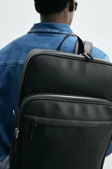 Mochila marrón de cuero sintético con textura grabada. Tiene un compartimento principal con cierre de cremallera, un bolsillo interno para dispositivos electrónicos de hasta 13 pulgadas y un bolsillo interno con cremallera. En la parte delantera tiene un bolsillo con cremallera. La parte posterior es acolchada y tiene una banda de sujeción para equipaje. Tiene un asa de mano y dos asas de hombro ajustables y acolchadas.