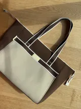 Bolso tipo tote de neopreno color marrón y beige, con cierre superior y asas de tela con detalles en marrón y blanco. Incluye monedero, bolsillo interno y externo con cierre, compartimentos internos y correa larga opcional.
