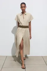 Vestido midi camisero color beige, confeccionado en lino. Presenta cuello solapa, mangas cortas con vuelta, jaretas delanteras, bolsillos laterales ocultos en costura, cierre frontal con botones y cinturón trenzado marrón.