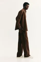 Camisa de corte oversized en denim de algodón rígido revestido, color marrón oscuro. Presenta cuello inglés, tapeta clásica con botones de metal y canesú en la espalda. Tiene hombros caídos, mangas largas con puños abotonados y bajo redondeado.