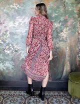 Vestido camisero midi de gasa estampada con flores rojas y blancas sobre fondo marrón. Tiene cuello con solapa, abertura central con botones, mangas largas y lazo de ajuste en la cintura.