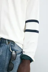 Camiseta blanca de manga larga con cuello en pico. Tiene el número 12 bordado en el pecho y dos franjas horizontales en color azul marino en cada manga.