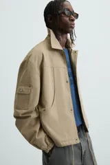 Cazadora color arena de corte relaxed fit, confeccionada en tejido de algodón. Cuenta con cuello solapa, manga larga con bolsillo multifunción, bolsillos frontales de plastrón, bolsillo interior, acabados con elástico y bajo ajustable con cordón. Cierre frontal con cremallera oculta por solapa con botones a presión.