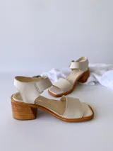 Sandalias de cuero color crudo, con tira ancha sobre los dedos, pulsera al tobillo con hebilla metálica y taco de madera de 6 cm de altura.