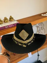 Sombrero de ala ancha tejido en hilo de coco y sintético, color marrón claro. La copa está cubierta con encaje naranja quemado con patrón floral. Tiene una cinta de cuero marrón oscuro y un aplique decorativo de hoja de gamuza con incrustaciones de pedrería.