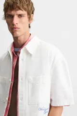 Camisa blanca de algodón con estructura canvas, corte boxy fit, cuello bowling y manga corta. Presenta bordado en azul claro con texto cursivo en el pecho y botones frontales.