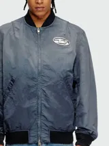 Campera bomber Diesel modelo J-Kepes, color gris azulado con efecto desgastado o tie-dye sutil. Presenta cuello, puños y cintura de punto elástico en color negro. Lleva un parche grande con el logo 'D' de Diesel en el pecho izquierdo.