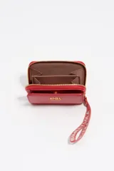 Billetera de cuero vacuno color rojo con estructura rígida y cierre de cremallera. Presenta un asa de tejido con el logo de la marca y el nombre de la firma en letras metálicas doradas en el frente.