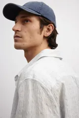 Camisa blanca de popelina de algodón texturizado, con cuello inglés, tapeta lisa, canesú con pliegue de caja en la espalda y mangas largas con abertura y cierre ajustable de botón en los puños. Corte holgado y bajo levemente redondeado.