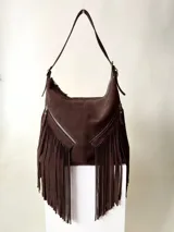 Bolso tipo hobo de cuero color marrón chocolate con flecos largos en la parte inferior y dos bolsillos frontales con cierre. Tiene correa de hombro ajustable y cierre principal con cremallera metálica YKK.