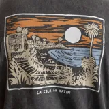 Remera negra de algodón con estampa en el pecho de una casa de surf, una palmera y el texto "La Isla by Katin".