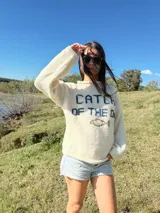 Sweater color crema tejido en lana merino, con cuello redondo y mangas largas. Presenta la frase "CATCH THE DAY" en color azul y un dibujo de un pez en el centro.