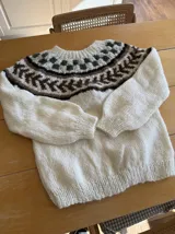 Sweater tejido a mano en lana mohair color crudo, con cuello redondo y mangas largas. Presenta un diseño con motivos geométricos en tonos marrón y verde en la parte superior del pecho.