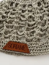 Gorro tipo beanie tejido a mano en hilo de algodón, color beige jaspeado, con diseño de malla calada y etiqueta de cuero marrón con el nombre 'Soriame' grabado.