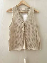 Chaleco de hilo de algodón color beige, tejido con punto calado y lurex. Tiene escote en V profundo y cordones tejidos que se entrelazan en el frente para atar. El corte es recto y suelto.