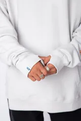 Buzo canguro gris claro de corte oversize, con capucha y cordones negros con letras blancas. Presenta un bolsillo tipo canguro y un estampado pequeño en el pecho y otro en la manga.