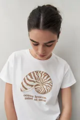 Remera blanca de manga corta con estampado de una concha marina y la frase "ocean breeze, mind at ease" en color marrón.