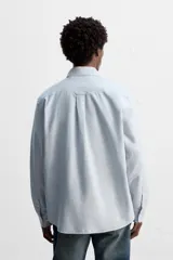 Camisa celeste a rayas verticales, de corte regular fit, confeccionada en tejido de algodón. Tiene cuello abotonado, manga larga con puños abotonados, bolsillo de parche en el pecho y cierre frontal con botones.