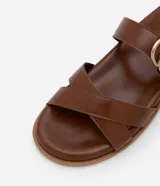 Sandalia plana marrón de cuero sintético, con punta redonda, tiras cruzadas y hebilla dorada lateral.