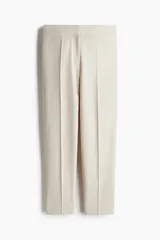 Pantalón cigarette color beige de tiro alto, con cierre y corchetes ocultos, bolsillos laterales discretos y bolsillos insertados decorativos atrás. Piernas ajustadas con abertura en los bajos y pliegues adelante y atrás.