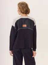 Buzo de manga larga con diseño de bloques de color y textura. El cuerpo principal es marrón, la espalda superior y hombros tienen un panel de tejido a rayas grises y azules, y los hombros superiores son de color beige rosado. Presenta costuras decorativas en rojo que separan los bloques de color.