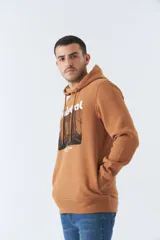 Buzo marrón con capucha y bolsillo canguro. Presenta un estampado frontal con el logo de la marca Mistral y una imagen de un arco arquitectónico. Confeccionado en algodón y poliéster.