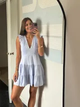 Vestido corto lila con escote en V y detalles de encaje o bordado en el busto y los hombros. Presenta un corte holgado con volados en la falda.