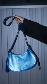 Cartera de hombro con forma de media luna, confeccionada en cuero vacuno con acabado metalizado azul brillante. Presenta manijas con ojales metálicos de color níquel y cierre de cremallera.