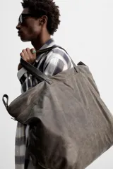Bolso de viaje marrón oscuro fabricado en piel con acabado craquelado. Tiene un compartimento principal con cierre de cremallera, un bolsillo plano interior con cremallera y dos bolsillos frontales con botón a presión. Cuenta con asas de mano de piel y asa de hombro removible y ajustable.