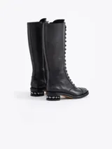 Bota de caña alta color negro, confeccionada en cuero vacuno, con cordones frontales marrones y cierre lateral. Presenta un diseño picado en la capellada y un taco de 4 cm con detalle de esferas metálicas.