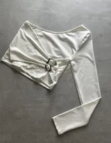 Top blanco de manga larga con corte irregular y abertura en el frente unida por una hebilla metálica con forma de corazón.