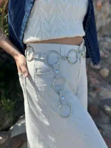 Cinto de cuero sintético color beige con tres aros metálicos plateados colgando.