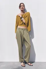 Pantalón color verde oliva de tiro medio con cintura con trabillas, bolsillos laterales, pinzas delanteras y raya planchada. Cierre frontal con cremallera, botón y ganchos metálicos.