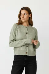 Cardigan de punto acrílico color verde claro, de corte corto y ajustado al cuerpo, con cuello redondo a la base y cierre frontal de botones marrones. Presenta trenzas decorativas en las mangas raglán.