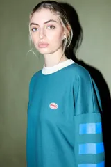 Buzo verde oscuro con cuello redondo y puños y dobladillo en color crema. Presenta un parche ovalado con el texto "CORAL" en el pecho y detalles de franjas azules en las mangas.