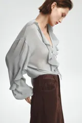 Blusa color marrón con escote en pico, manga larga abullonada con elástico en los puños y volantes en el escote y mangas.