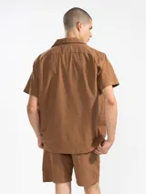 Camisa azul de hombre de calce relajado y corte cuadrado, confeccionada en una mezcla liviana de lino y algodón. Presenta cuello tipo camp, cierre con botones de madera grabados con el logo y un bordado sutil en el pecho izquierdo con forma de palmera. Incluye patch con logo de la marca.