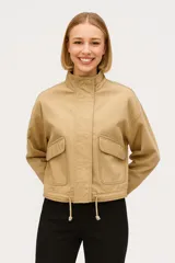 Campera corta color beige, de corte holgado, con cuello alto, cierre frontal con cremallera, bolsillos con solapa y cordón ajustable en el ruedo.