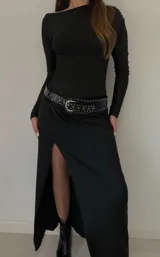 Vestido largo negro de mangas largas, con escote redondo y tajo lateral. Incluye cinturón de cuero negro con hebilla plateada y tachas.