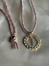 Collar con dije circular dorado con strass y letra 'A' en el centro, con cordón trenzado multicolor y cierre ajustable.