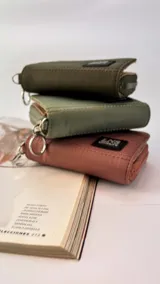 Billetera corta con cierre alrededor y tapa, confeccionada en tela capitoneada. Incluye varias reparticiones con tarjeteros y bolsillos internos, y una correa de mano desmontable. Disponible en negro, rojo y dorado.