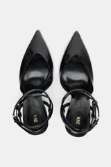 Zapato de tacón destalonado color negro charol, con punta fina, tacón fino alto y tiras finas con hebillas ajustables al tobillo.