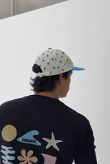 Gorro tipo jockey color beige con lunares azules, visera azul y bordado frontal con la palabra "ABOIU" en letras de colores y estrellas.