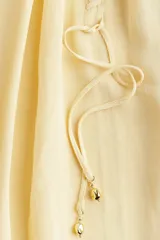 Blusa color crema de gasa transparente con cuello elevado y mangas globo largas con puños elásticos. Presenta pespuntes sobredimensionados decorativos en las costuras raglán y tira diagonal adelante con colgantes de cuentas brillantes.